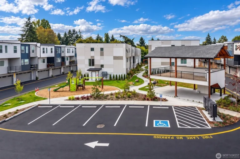 Exterior view of a modern townhouse complex featuring a playground, winding walking path, covered pavilion, and a parking lot under a blue sky with clouds
