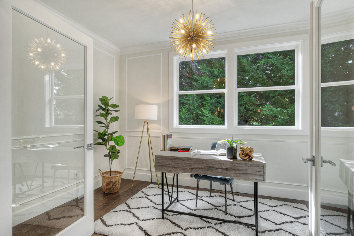 A modern home office with a minimalist desk, a decorative light fixture, and large windows overlooking lush greenery, ideal for remote work.