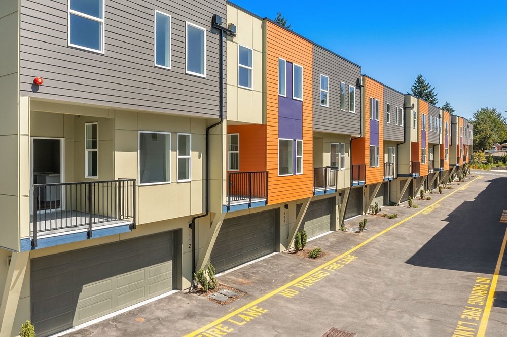 A row of newly built modern townhomes with colorful siding, showcasing the exterior and garage design of a TP Home Builds community in Washington.