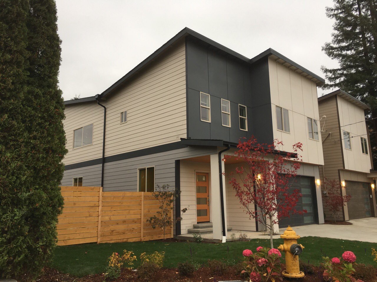 A modern two-story home with a blend of beige and dark gray siding, featuring a wood-panel garage door and a landscaped front yard.