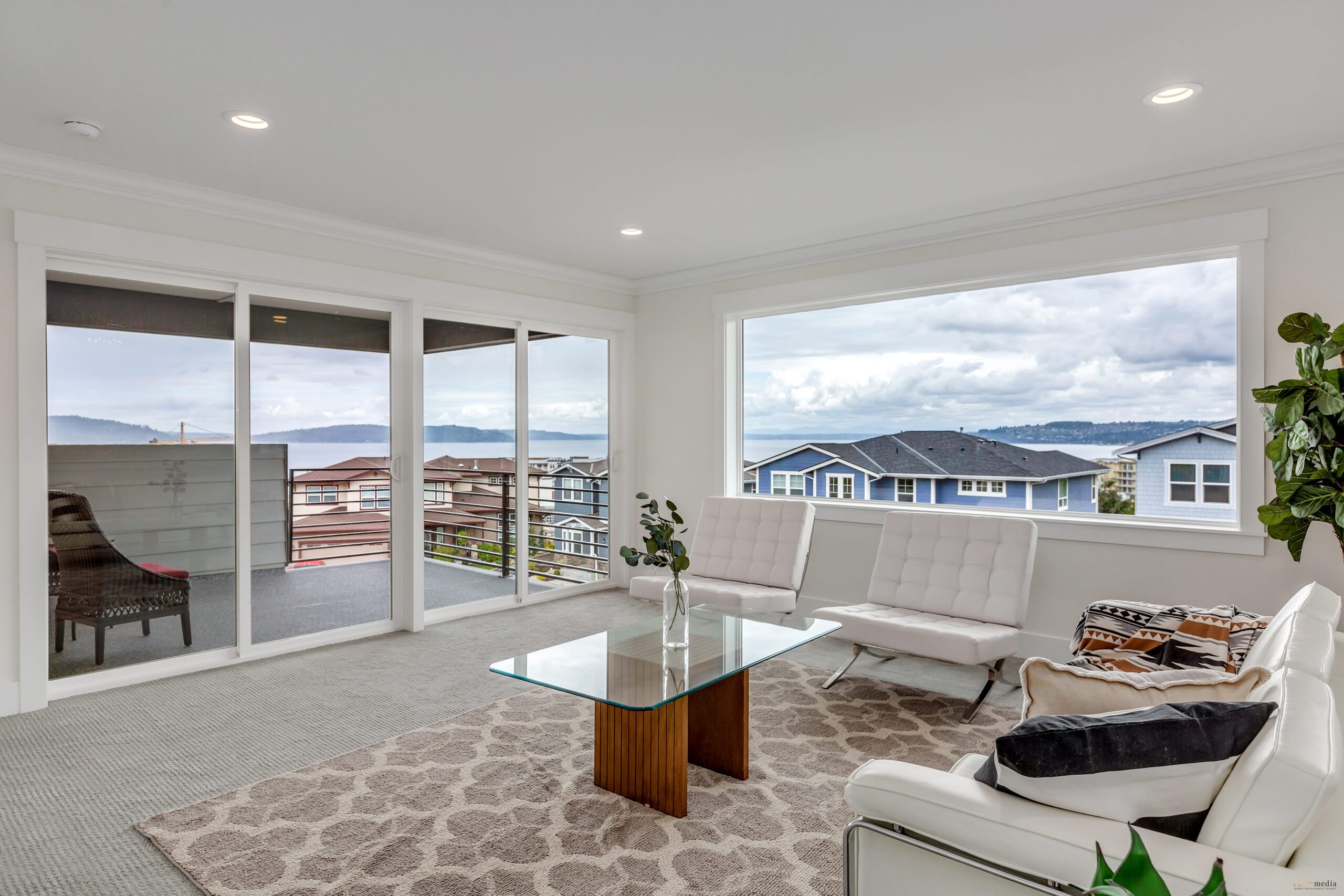 A modern living room with sliding glass doors leading to a balcony, offering a panoramic view of the water and a nearby neighborhood.