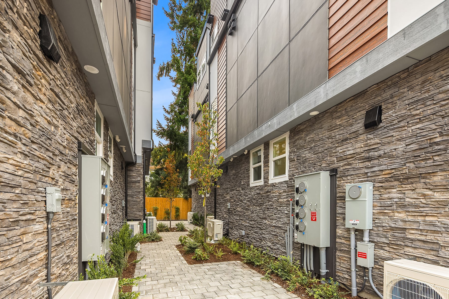 A narrow alleyway between two homes, featuring a stacked stone facade and a brick paver walkway, highlighting the unique exterior finishes of an ADHG development.