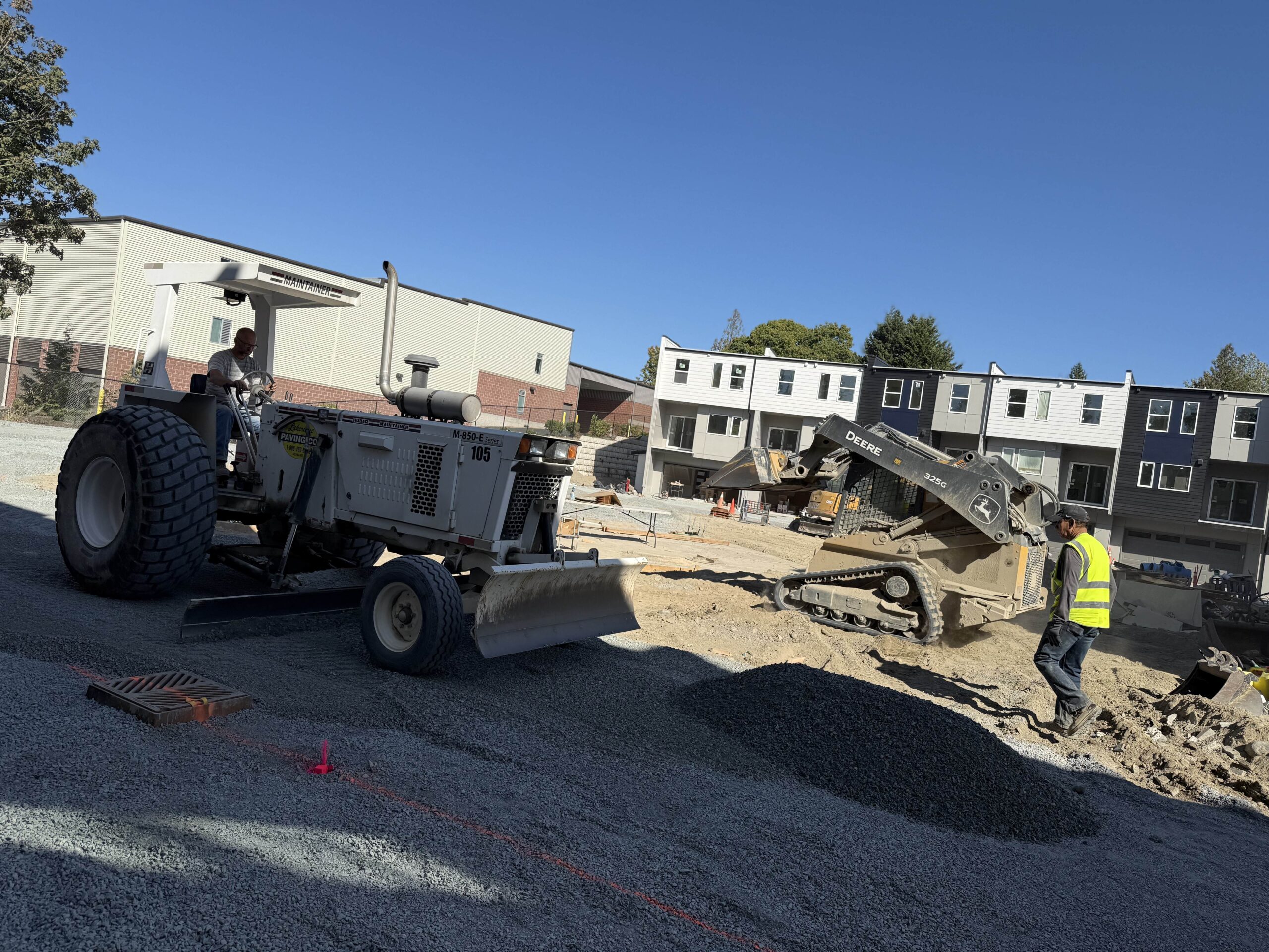 Large grading equipment and a small excavator work to level a gravel surface, showing the earthwork and site preparation phase of construction.